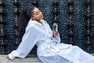 A relaxed woman in a white robe enjoys a glass of sparkling drink, sitting beside a wall with cascading water and smooth stones.