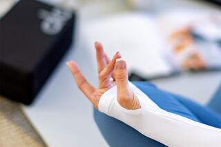 A relaxed hand forms a meditation gesture, wearing a white long-sleeve shirt. A yoga mat and magazines are in the background.