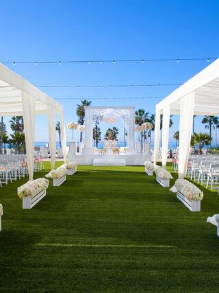 A beautifully decorated outdoor wedding venue features white drapes, floral arrangements, and a serene view of palm trees and blue skies.