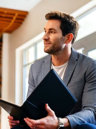A young man in a stylish blazer sits with a laptop, looking thoughtfully towards the side, in a bright, modern interior.