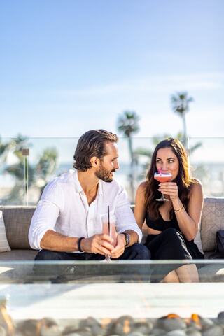 A glass of white wine stands next to a gourmet dish on a table, with a blurred beach view and a cocktail in the background.