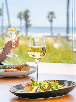 A glass of white wine stands next to a gourmet dish on a table, with a blurred beach view and a cocktail in the background.