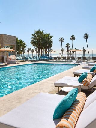 A bright, inviting pool area lined with lounge chairs, surrounded by palm trees and sun umbrellas, overlooking the ocean on a clear day.