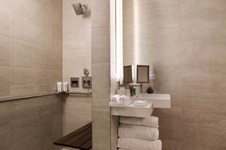 A modern, minimalist bathroom features beige tiles, a wooden bench, sleek fixtures, and neatly arranged towels and toiletries.