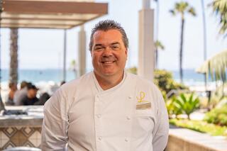 A smiling chef in a white uniform stands outdoors by the beach, with palm trees and a sunny atmosphere in the background.
