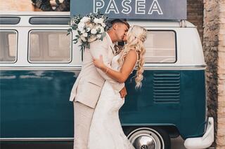 A joyful couple kisses in front of a vintage blue van, surrounded by wedding decorations and holding a bouquet, celebrating their love.
