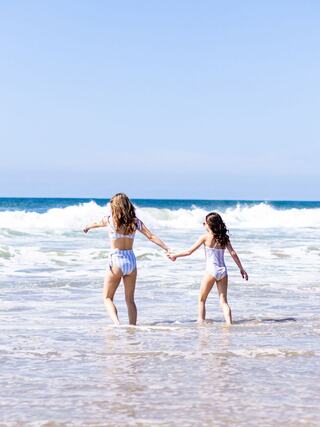 Two girls walk hand in hand into shimmering waves, enjoying a sunny beach day with clear blue skies and gentle surf.